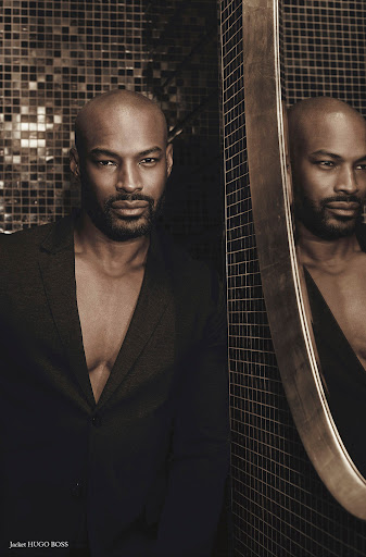 Bald Black man with trimmed beard wearing a black jacket next to a round mirror