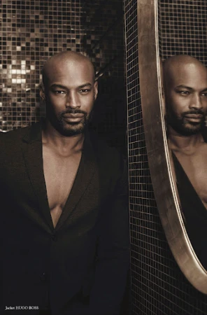Bald Black man with trimmed beard wearing a black jacket next to a round mirror