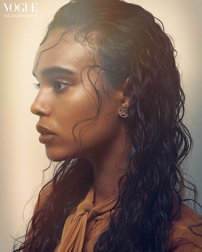 Profile portrait of a woman with dark, curly, wet hair and tan skin. She wears a tan top and has gold earrings in front of a blurred background.