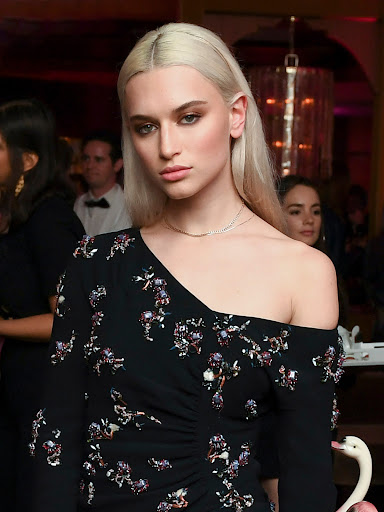 Elegant woman with platinum blonde hair poses in a black floral dress indoors, attending a party with a blurred background.
