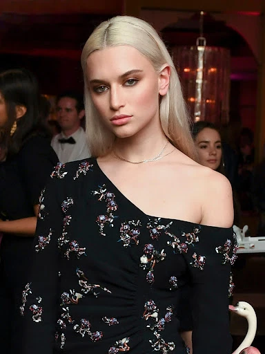 Elegant woman with platinum blonde hair poses in a black floral dress indoors, attending a party with a blurred background.