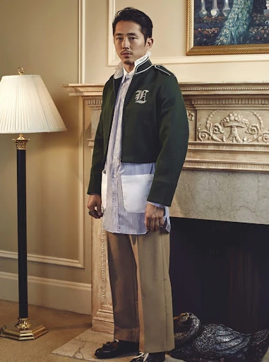 Adult male in fashion shot, wearing dark green jacket, striped button-down shirt, tan pants, standing inside by a fireplace mantle.