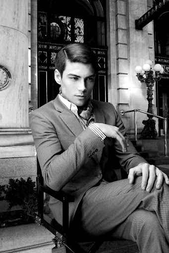Black and white shot of a man in a suit sitting on a director's chair in front of a building with marble columns and a streetlamp.