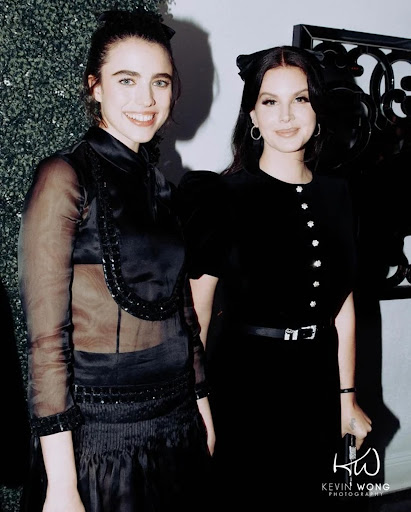 Two women in elegant black dresses pose indoors. One wears a sheer-sleeved dress, while the other has a cat-ear headband and buttoned dress.
