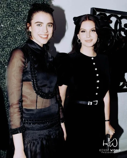 Two women in elegant black dresses pose indoors. One wears a sheer-sleeved dress, while the other has a cat-ear headband and buttoned dress.