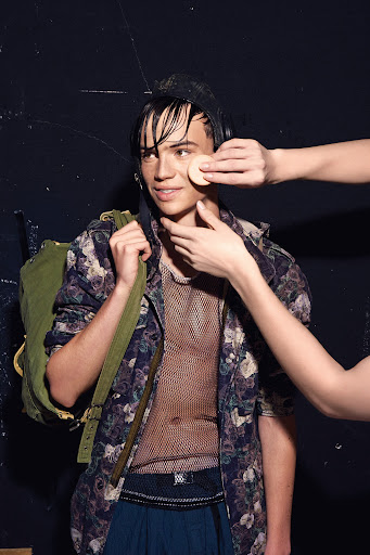A young male model having makeup applied backstage by two hands with a sponge, wearing a floral patterned jacket and mesh top.