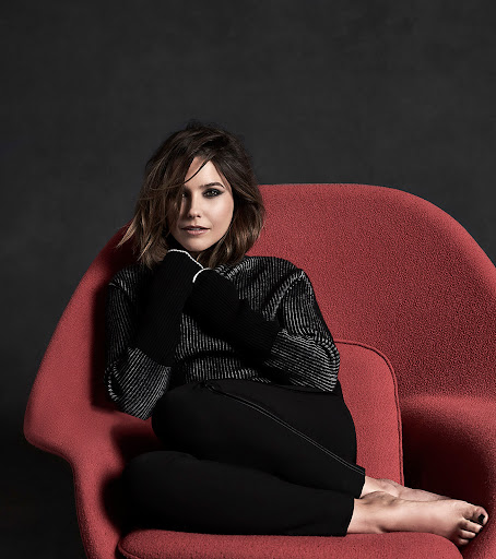 Adult woman with short brown hair sits in a red chair with her legs crossed and her arms in her lap in a photo studio.