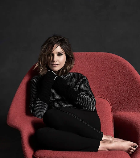 Adult woman with short brown hair sits in a red chair with her legs crossed and her arms in her lap in a photo studio.