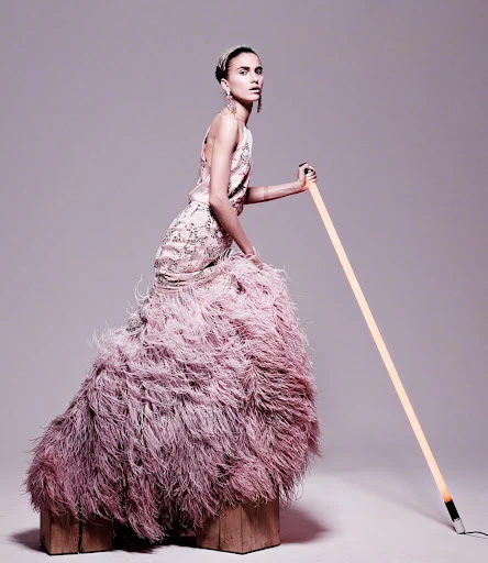 Elegant woman in feathered gown poses with a glowing light tube in a studio. Fashion shoot showcasing couture and creative lighting.