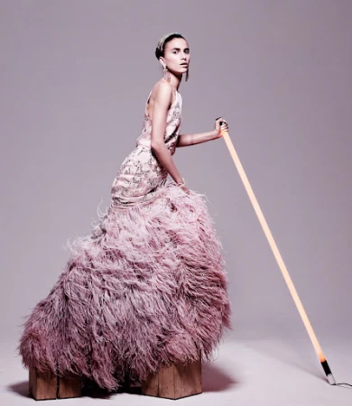 Elegant woman in feathered gown poses with a glowing light tube in a studio. Fashion shoot showcasing couture and creative lighting.