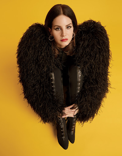A woman poses in black boots and a feathered jacket against a yellow backdrop, her hands resting on her boots in a studio setting.