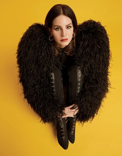 A woman poses in black boots and a feathered jacket against a yellow backdrop, her hands resting on her boots in a studio setting.