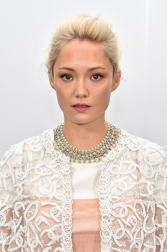 Portrait of a woman with blonde hair wearing a white embroidered jacket and heavy pearl statement necklace