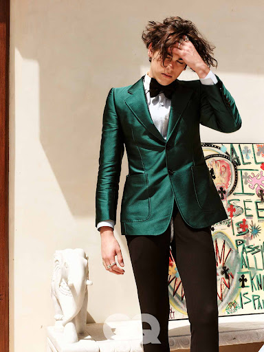 Stylish young man in a green suit and bow tie poses indoors with an abstract painting and a decorative elephant statue.
