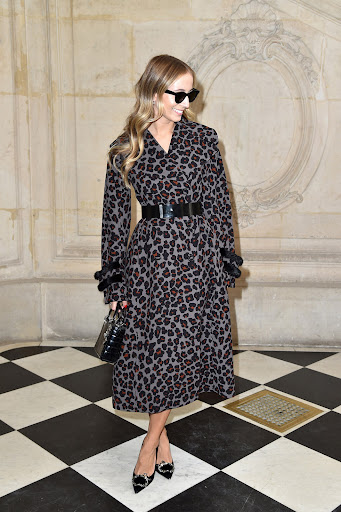 Blonde woman in a leopard print coat, black belt, and black heels stands in a marble lobby with a checkered black and white floor.