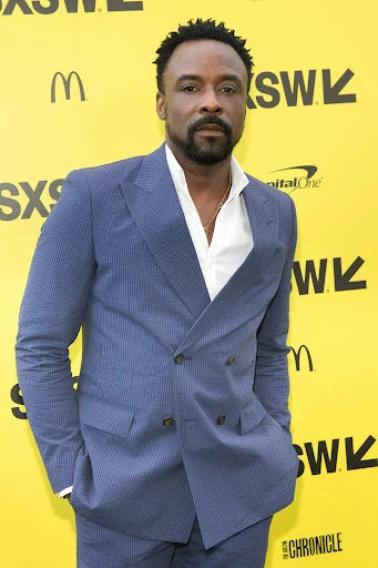Portrait of a dark-skinned man in a blue suit at SXSW with a yellow background, exuding confidence and professionalism at an entertainment event.