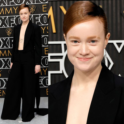 Smiling woman in black suit poses on red carpet at an awards show; sleek auburn hair and geometric silver earrings.