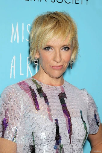 Blonde woman with short hair, wearing a patterned dress, at a premiere event against an aqua background with event signage.