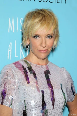Blonde woman with short hair, wearing a patterned dress, at a premiere event against an aqua background with event signage.