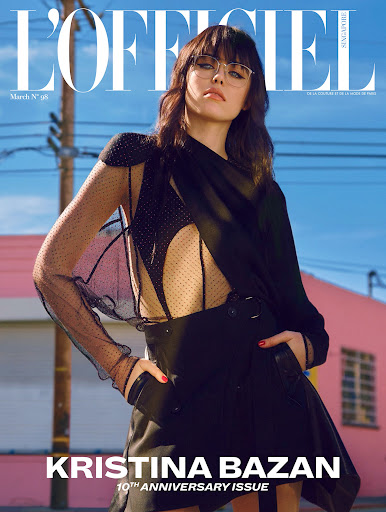 Fashion magazine cover featuring a woman with bangs wearing glasses and a black dress against a blue sky and pink building backdrop.