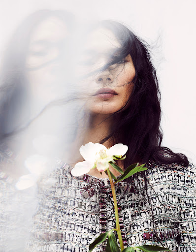 Portrait of an adult woman with long dark hair wearing a patterned tweed jacket, holding a white flower against a white background.