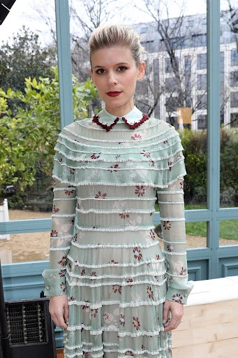 A fair-skinned woman with blonde hair and dark red lipstick wears a light green dress with layers and a vintage collar outdoors.