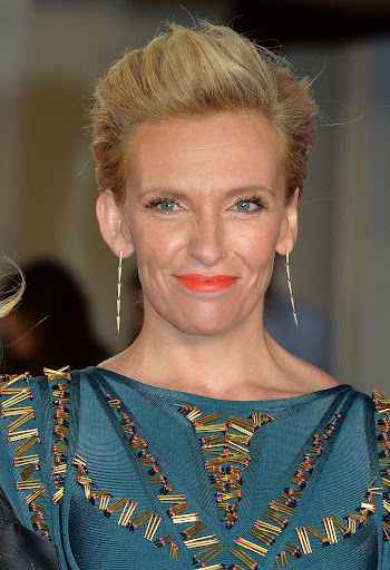 Close-up of Toni Collette on a red carpet wearing a teal dress with gold accents, diamond earrings, and bright coral lipstick.