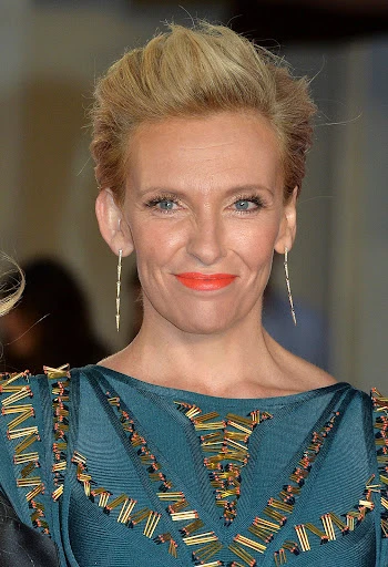 Close-up of Toni Collette on a red carpet wearing a teal dress with gold accents, diamond earrings, and bright coral lipstick.