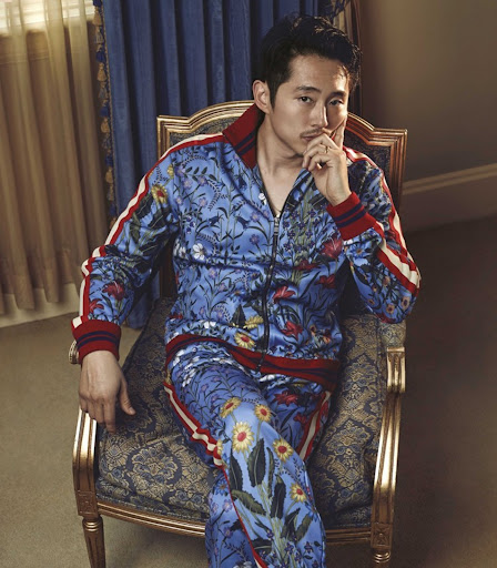 Portrait of an Asian man with dark hair, wearing a blue floral tracksuit, sitting on a chair with gold accents in an indoor setting.