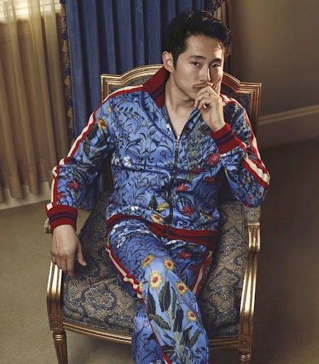 Portrait of an Asian man with dark hair, wearing a blue floral tracksuit, sitting on a chair with gold accents in an indoor setting.