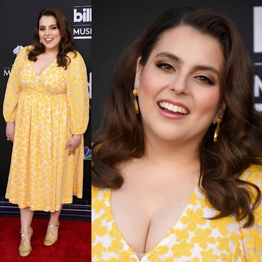 Beanie Feldstein poses in a yellow floral dress on a red carpet at Billboard Music Awards. She has shoulder-length curly brown hair.