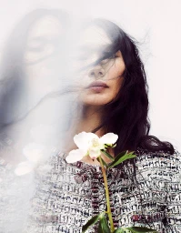 Portrait of a woman with long dark hair holding a white flower in a patterned tweed jacket with a blurred overlay effect.