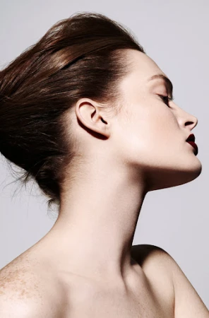 Profile portrait of a woman with dark red lips, styled hair, and freckles against a neutral background.
