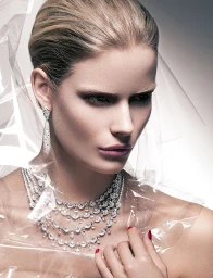 Close-up portrait of a woman wearing layered diamond necklaces and drop earrings behind a translucent plastic sheet.