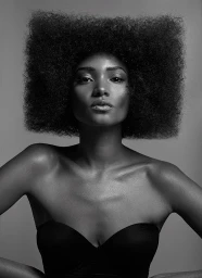Portrait of a woman with a large geometric square afro hairstyle wearing a black strapless top against a grey background