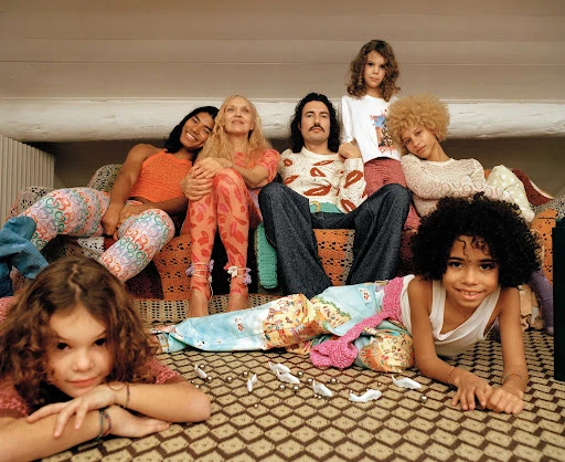A family portrait shows two adult women, one adult man, and four children sitting and lying on an ornate, vintage-style couch and carpet.