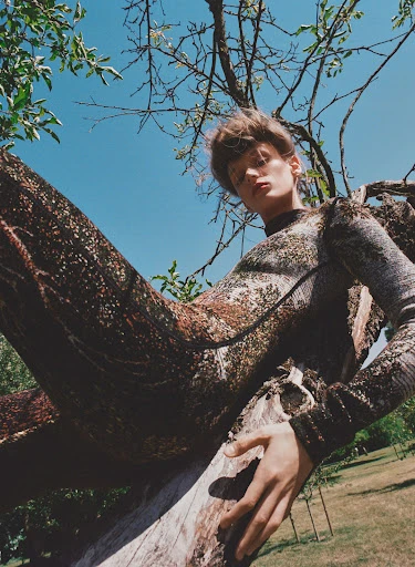 Adult woman wearing patterned jumpsuit poses on tree branch against blue sky in nature. Fashion editorial photography in outdoor setting.