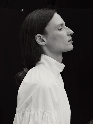 Black and white profile portrait of a model with dark hair tied back in a low ponytail, wearing a white high-necked shirt.