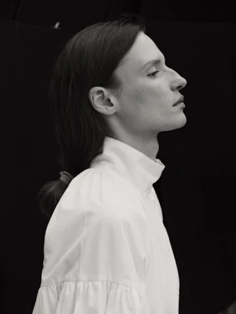 Black and white profile portrait of a model with dark hair tied back in a low ponytail, wearing a white high-necked shirt.