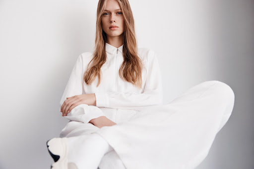 A woman with long blonde hair in a white outfit poses in a studio, looking directly at the camera with a neutral expression.