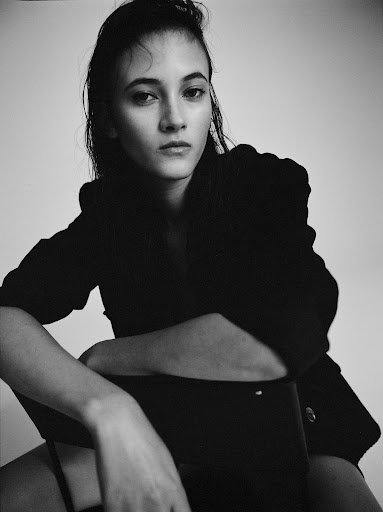 Portrait of a woman with dark wet-look hair sitting backward on a chair wearing a dark blazer.
