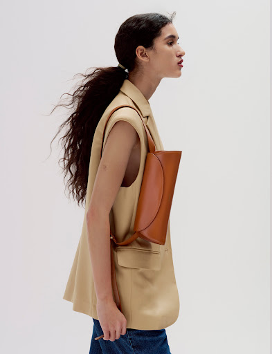 A woman wearing a tan blazer and blue jeans is walking, carrying a brown handbag in this fashion-oriented studio shot on a white backdrop.