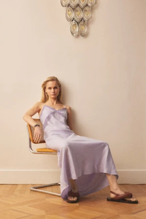 Blonde woman in lilac dress poses sitting on a chair in a neutral setting with parquet floor and modern glass lighting fixture.