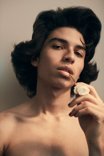 Portrait of a young man with long dark hair holding a white rose in his mouth in a studio setting with neutral lighting.