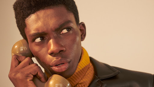Close-up portrait of an adult male with dark skin holding a vintage telephone to his ear, wearing a brown leather jacket.
