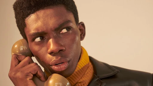 Close-up portrait of an adult male with dark skin holding a vintage telephone to his ear, wearing a brown leather jacket.