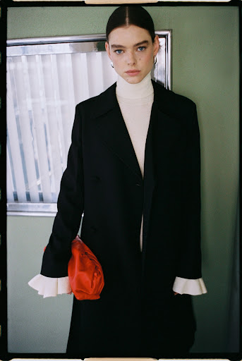 Young woman in a black coat and cream turtleneck poses indoors in front of a muted green wall with white molding and window detail.