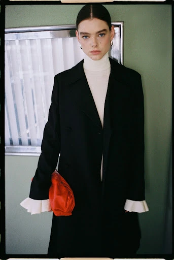 Young woman in a black coat and cream turtleneck poses indoors in front of a muted green wall with white molding and window detail.