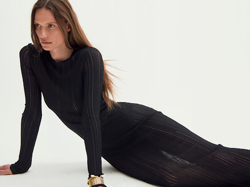 Woman in a long black ribbed dress posing against a plain white studio background with long brown hair