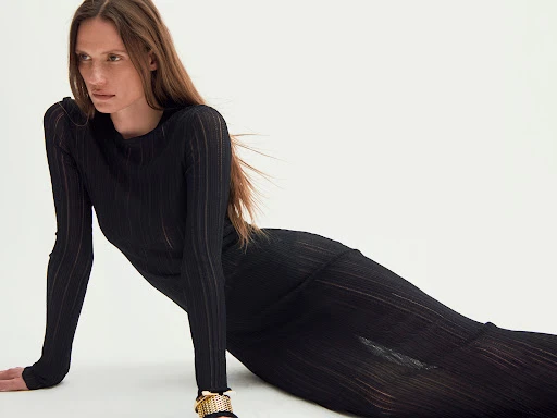Woman in a long black ribbed dress posing against a plain white studio background with long brown hair